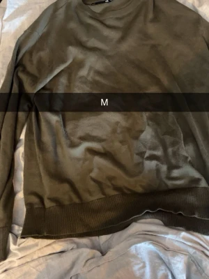 Olivgrön stickad sweatshirt i storlek M - En basic olivgrön stickad sweatshirt med rund hals och ribbade muddar vid ärmslut och nederkant. Perfekt för en avslappnad stil och enkel att matcha med jeans eller joggers.