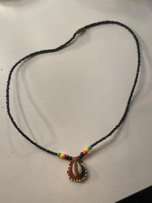 Halsband med snäcka och färgglada pärlor - Unikt halsband med svarta små pärlor och färgglada detaljer i rött, gult, grönt och turkost. Hängsmycket är en snäcka infattad i brunt material med dekorativa sömmar. Perfekt accessoar för en bohemisk stil. Helt oanvänt 🫶