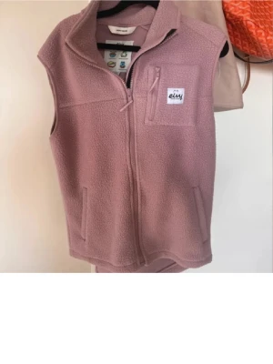 Rosa teddyväst från Eivy - Mysig rosa fleeceväst från Eivy med dragkedja framtill, två sidofickor och en bröstficka med logotyp. Perfekt att slänga på sig över en hoodie eller tröja för extra värme. Enkel och stilren design med hög krage.