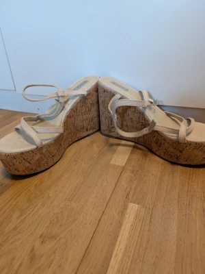 Beige platåsandaler med korksula - Säljer ett par snygga beige platåsandaler från Bianco med höga kilklackar i kork. Sandalerna har tunna remmar över foten och runt ankeln för en trendig look. Perfekta för dig som vill ha extra höjd och en somrig vibe.