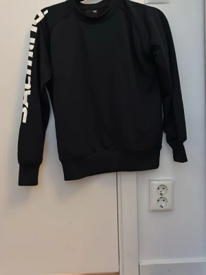 Svart träningströja från Salming - Cool svart träningströja från Salming med vit logga längs ena ärmen. Tröjan har rund halsringning, ribbade muddar och är tillverkad i ett mjukt syntetmaterial. Perfekt för en sportig och avslappnad stil.