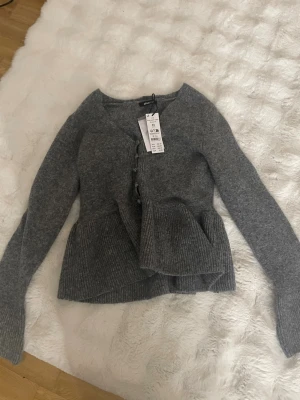 Grå kofta med volang från Gina Tricot - Superfin grå kofta från Gina Tricot med volangdetalj nedtill och knäppning framtill. Långärmad och stickad i mjukt material, perfekt för lager-på-lager. Enkel att styla till jeans eller kjol.