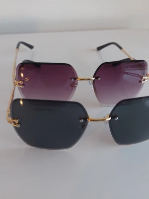 Två par kantiga solglasögon guld - Säljer två par trendiga solglasögon med kantiga glas och guldfärgade bågar. Ett par har mörkgrå glas och det andra har lila tonade glas. Båda har minimalistisk design utan synlig ram runt glasen och svarta skalmar. Perfekta för att lyfta din stil i sommar.
