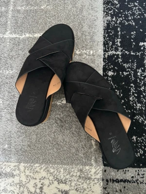 Svarta slip-in sandaler med korsade band - Snygga svarta sandaler med breda korsade band i mockaliknande material. Slip-in modell med öppen tå och innersula i beige. Köpta för 350kr hos DinSko