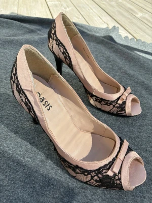 Rosa spetsklädda pumps från Oasis - Snygga pumps i ljusrosa tyg från Oasis med svart spetsdetalj över hela skon. Modellen har öppen tå och en liten rosett framtill för extra stilpoäng. Klacken är smal och elegant, perfekt för att lyfta vilken outfit som helst.