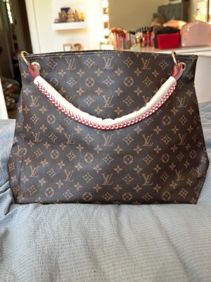 Louis Vuitton Monogram handväska - Snygg handväska från Louis Vuitton i klassiskt brunt monogrammönster. Väskan har en vit handtag med röd detaljsöm och guldiga metalldelar. Tillverkad i canvas med läderdetaljer, perfekt för dig som gillar lyxiga accessoarer. Har inget äkthetsbevis därav pris!