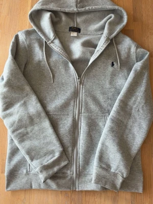 Grå hoodie från Polo Ralph Lauren - Snygg grå zip-hoodie från Polo Ralph Lauren med klassisk huva och dragsko. Broderad logga på bröstet och två fickor framtill. Mjuk bomullsblandning som är perfekt för en chill och avslappnad stil.