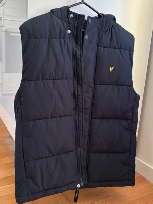 Svart dunväst från Lyle & Scott - Snygg svart dunväst från Lyle & Scott med huva och dragkedja framtill.                                               ‼️Original pris på butik: 1000kr‼️