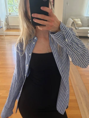 Blåvit randig skjorta i Linné - Snygg blå skjorta med vita ränder och klassisk krage. Skjortan har långa ärmar och knäppning framtill. Tillverkad i mjuk bomull, perfekt för en avslappnad men stilren look.