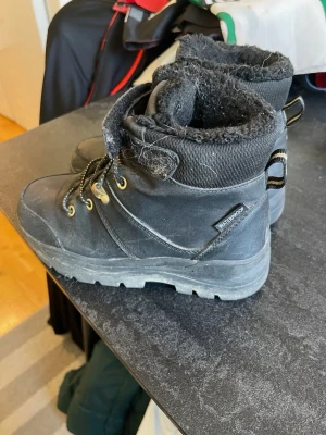 Svarta vinterkängor med foder stl 33 - Svarta kängor från Zoey med grov sula, snörning och kardborreband upptill. Utsidan är i slitstarkt material och insidan har mjukt, fluffigt foder för extra värme. Metallöglor i guldfärg ger en snygg kontrast. Märkta med 'Waterproof' på sidan.