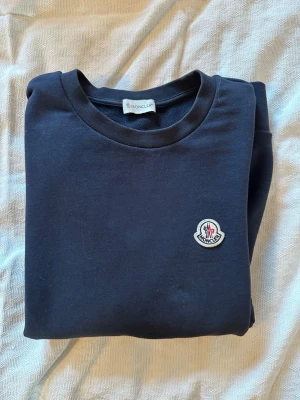 Svart sweatshirt från Moncler - Snygg svart sweatshirt från Moncler med rund halsringning och klassisk logotyp på bröstet. Tröjan har en enkel och stilren design, perfekt för en avslappnad look. Tillverkad i mjukt material för extra komfort.