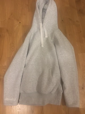 Ljusgrå hoodie från Russelious - Ljusgrå hoodie från Russelious med klassisk känguruficka och ribbade muddar. Tröjan har en stor huva med dragsko och broderad logga på ärmen. Perfekt för en avslappnad och stilren look.