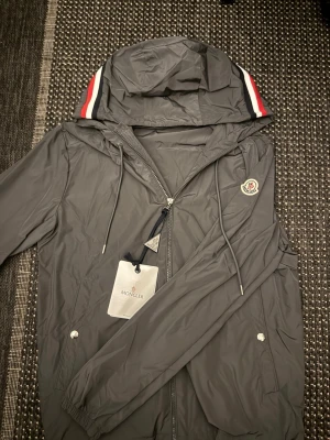 Moncler jacka - Snygg grå vindjacka från Moncler med huva och dragkedja framtill. Jackan har Moncler-logga på bröstet och röda, vita och svarta ränder på huvan. Justerbara snören vid huva och midja samt fickor med tryckknappar. Perfekt för blåsiga dagar.