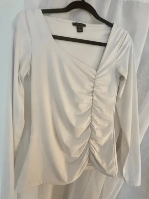 Vit rynkad topp - Snygg vit tröja/topp med lång ärm och rynkdetalj framtill. Toppen har en v-ringad hals och är tillverkad i mjukt, stretchigt material som sitter bekvämt på kroppen. Perfekt för dig som gillar stilrena plagg med en twist. 