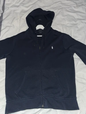 Svart hoodie från Polo Ralph Lauren - Snygg svart hoodie från Polo Ralph Lauren med dragkedja, huva med dragsko och klassisk vit logga på bröstet. Mjuk bomullsblandning och två stora fickor framtill. Perfekt för en chill och stilren look.