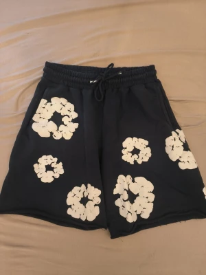 Denim tears mörkblå shorts  - Säljer ett par mörkblå Denim tear med vita blommiga tryck och snörning i midjan. De har en avslappnad passform och elastisk midja, perfekta för sommarens chill dagar. Materialet känns mjukt och bekvämt, det är bommul
