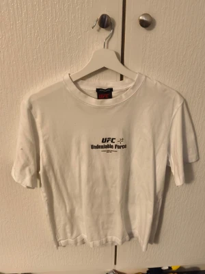 Vit UFC t-shirt med tryck - Vit t-shirt från UFC med tryck på både fram- och baksidan. Framsidan har texten 'UFC Undeniable Force' och baksidan har stort UFC-tryck och texten 'Ultimate Fighting Championship'. Klassisk passform och rund hals. Perfekt för dig som gillar kampsport och streetwear.