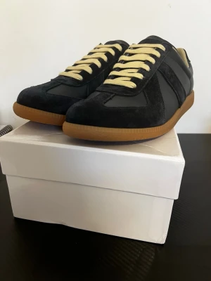 Maison Margiela skor  - Snygga svarta sneakers från Maison Margiela med beige snörning och klassisk gummisula. Skorna har en mix av mocka och skinn, vilket ger en lyxig känsla. Perfekta för dig som gillar stilrena och trendiga sneakers med en retrovibe. Av märket Maison Margiela, riktigt fina skor inför sommaren 
