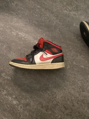Nike Air Jordan 1 Mid röd/svart/vit - Säljer ett par Nike Air Jordan 1 Mid sneakers i klassisk färgkombination av rött, svart och vitt. Skorna har svart läder på tå och häl, röd tåbox och plös, samt vit sidopanel med röd swoosh. Ikoniska Air Jordan-loggan på sidan och svart snörning.