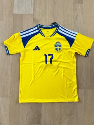 Svensk gyökeres landslagströja VM 2026 - Gul svensk landslagströja från Adidas med blå detaljer, nummer 17 på bröstet och det svenska fotbollsförbundets emblem. Tröjan har korta ärmar, rund hals och klassiska Adidas-ränder på axlarna. Tillverkad i lätt och ventilerande material, perfekt för fotboll.