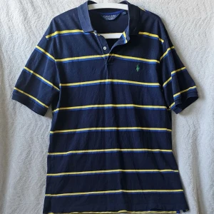 Randig pikétröja från Polo Ralph Lauren - Mörkblå pikétröja från Polo Ralph Lauren med gula och blå ränder. Klassisk krage, korta ärmar och broderad logga på bröstet. Perfekt för en sportig och avslappnad stil.