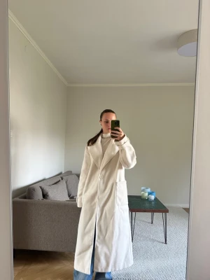 Lång cremevit kappa med bred krage - Snygg lång kappa i cremevitt med breda slag och knäppning framtill. Kappan har raka ärmar, två stora fickor och en stilren, minimalistisk look. Perfekt för dig som gillar en clean och trendig stil.