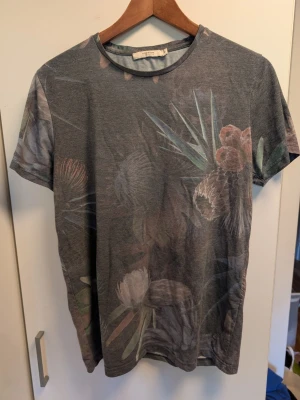 Grå blommig t-shirt från Jack & Jones - Snygg grå t-shirt från Jack & Jones Premium med diskret blommigt mönster i dova färger. T-shirten har rund halsringning och korta ärmar, perfekt för en avslappnad stil. Tillverkad i mjuk bomull för skön komfort.