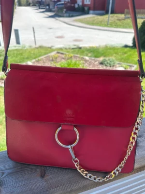 Röd axelväska från Chloé - Snygg röd axelväska från Chloé med guldfärgad kedja och ringdetalj framtill. Väskan har flera fack på insidan och en stilren, fyrkantig form. Materialet är slätt och mockaliknande läder. Perfekt för dig som vill ha en statementväska med lyxig känsla.
