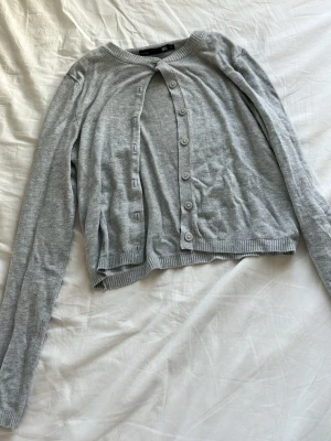 Ljusgrå cardigan från lager 157 - Ljusgrå cardigan med rund halsringning och långa ärmar från lager 157. Tröjan har knappar framtill och ribbade muddar vid ärmslut och nederkant. Perfekt att slänga över en topp för en enkel och clean look.