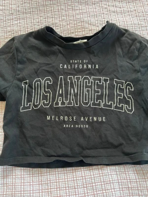 Svart croppad Los Angeles t-shirt - Svart croppad t-shirt med tryck 'Los Angeles' och 'Melrose Avenue' framtill. T-shirten har korta ärmar och rund halsringning. Perfekt för en avslappnad streetstyle-look. Från Kappahl i storlek 134/140