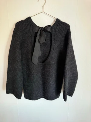 Svart stickad tröja med rosett - Mysig svart stickad tröja med öppen rygg och en snygg rosett i nacken. Tröjan har lång ärm och är tillverkad i ett fluffigt, mjukt material som känns skönt mot huden. Perfekt för dig som gillar en stilren look med en twist.