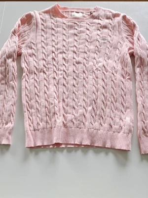 Ljusrosa kabelstickad tröja GANT - Säljer en jättefin ljusrosa kabelstickad tröja med rund halsringning. Ett tidlöst plagg som passar perfekt till både jeans och mer uppklädda tillfällen. Tröjan har en liten diskret logotyp broderad på bröstet.