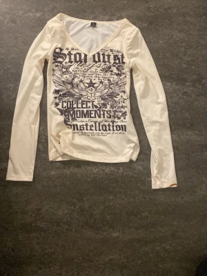 Vit långärmad topp med tryck - Cool vit långärmad topp med stort svart tryck framtill med text och grafiska detaljer. V-ringad hals och figurnära passform. Perfekt för dig som gillar statement-plagg och edgy stil.