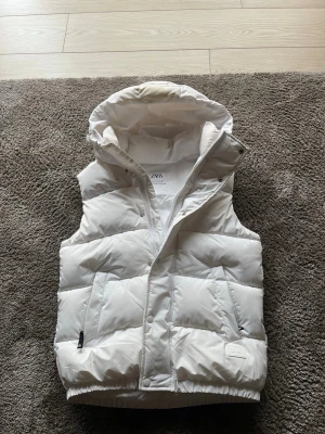 Vit dunväst från Zara med huva - Snygg vit dunväst från Zara med stor vadderad huva och dragkedja framtill. Västen har två fickor med dragkedja och quiltad design som ger en modern look. Perfekt för lager-på-lager-stil under kyliga dagar.