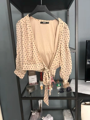 Beige omlottblus med svarta prickar - Trendig beige omlottblus från Bik Bok med svarta prickar och knytband i midjan. Blusen har långa puffärmar och är tillverkad i ett lätt, transparent tyg som ger en luftig känsla. Perfekt att styla med jeans eller kjol för en cool look.