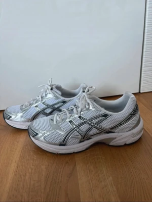 Vita Asics sneakers med silverdetaljer - Snygga vita Asics sneakers med mesh-ovandel och silverfärgade detaljer. Skorna har klassisk låg profil, vita skosnören och den ikoniska Asics-loggan på sidan. Perfekta för dig som gillar sportig och clean stil.