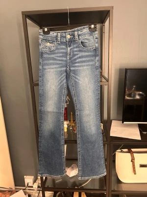 Blå bootcut jeans med broderade fickor - Snygga blå bootcut jeans med broderade detaljer på bakfickorna och klassisk femficksdesign. Jeansen har en ljus tvätt och subtila slitningar framtill. Perfekta för dig som gillar en avslappnad men trendig stil.