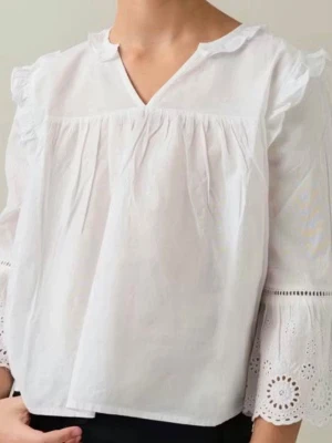 Vit blus med broderade volangärmar - Söt vit blus i bomull med v-ringning och volangdetaljer. Ärmarna är långa och avslutas med broderade spetsdetaljer som ger en romantisk känsla. Perfekt att styla med jeans eller kjol för en trendig look.