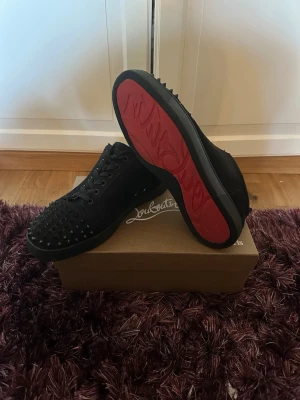 Svarta Louboutin sneakers med nitar - Snygga svarta sneakers från Christian Louboutin med klassisk röd sula och svarta nitar över tån. Skorna har lågt skaft, snörning och är tillverkade i mocka. Perfekta för dig som vill sticka ut med en edgy och lyxig stil. Storlek 45.