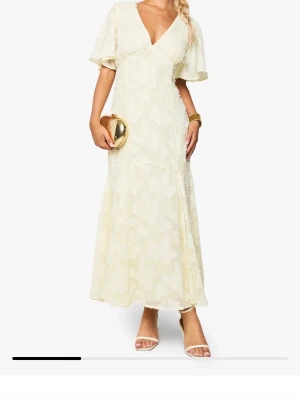 Ljus långklänning med volangärmar - Elegant ljus långklänning med v-ringning och korta volangärmar. Klänningen har ett diskret blommigt mönster och är tillverkad i ett lätt, flowy material som ger en romantisk känsla. Perfekt för sommarens speciella tillfällen.