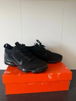 Nike Air Vapormax Flyknit  - Svarta Nike Air Vapormax Flyknit sneakers med stickad ovandel och genomskinlig sula. Swoosh-logga på sidan och snörning framtill. Lätta och flexibla med sportig look och modern design. Perfekta för dig som gillar streetwear och komfort.