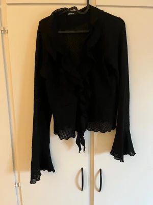 Svart volangkofta från Gina Tricot - Snygg svart kofta från Gina Tricot med volangkant längs öppningen och vid ärmslut. Koftan har knytning framtill och är tillverkad i ett tunt material. Perfekt för lager-på-lager och ger en cool touch till din outfit. Katt finns i hemmet!