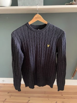 Mörkblå kabelstickad tröja Lyle & Scott - Snygg mörkblå kabelstickad tröja från Lyle & Scott med rund halsringning och den klassiska gula logon broderad på bröstet. Tröjan har långa ärmar och ribbade muddar vid ärmslut och nederkant. Perfekt för lager på lager och stilren vardagslook. Använd 1 gång som ny. Nypris 1000kr