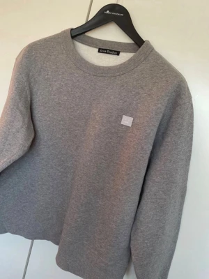Grå sweatshirt från Acne Studios - Säljer en stilren grå sweatshirt från Acne Studios med klassisk rund halsringning och diskret logopatch på bröstet. Tröjan är i mjukt material och har långa ärmar samt ribbade muddar vid ärmslut och nederkant.