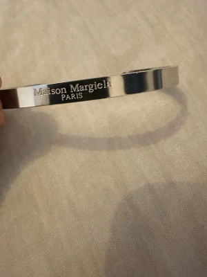 Maison Margiela silverfärgat armband - Snyggt och stilrent armband från Maison Margiela i silverfärgad metall. Armbandet är stelt och har en öppen design med ingraverad logotyp på utsidan. Perfekt accessoar för dig som gillar minimalistisk och modern stil.