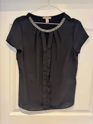 Svart blus med pärldetaljer Anna Field - Snygg svart blus från Anna Field med korta ärmar och rund halsringning dekorerad med pärlor och glittriga stenar. Blusen har ett lätt, tunt material och veck framtill för en elegant look. Perfekt att styla med jeans eller kjol.