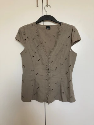 Grön blus med broderi från Gina Tricot - Söt grön blus med korta ärmar från Gina Tricot. Blusen har broderade detaljer, hålmönster och knappar framtill. V-ringad med spetskant längs urringningen och knappraden. Perfekt för en avslappnad och trendig look.