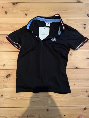 Svart pikétopp från Moncler - Svart pikétopp från Moncler med klassisk krage och korta ärmar. Kragen har blå insida och röda samt vita ränder längs kanterna. Moncler-logga broderad på bröstet. Perfekt för en sportig och stilren look.
