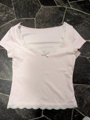 Ljusrosa topp med spetsdetaljer stl xs - Supersöt ljusrosa topp med korta ärmar, vit spetskant vid halsringning och nederkant samt en liten rosett framtill. Perfekt för en söt och feminin look. Mjuk och stretchig bomullskänsla.