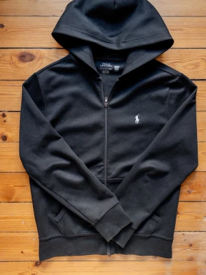 Svart hoodie från Polo Ralph Lauren - Svart hoodie med dragkedja från Polo Ralph Lauren. Klassisk design med vit broderad logga på bröstet, två fickor fram och ribbade muddar. Tillverkad i mjukt bomullsmaterial och har en stor huva för extra komfort.